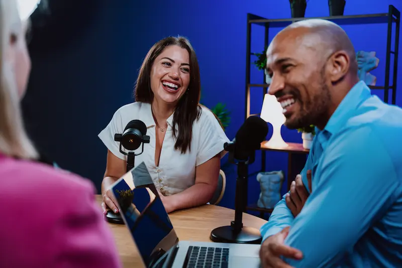 Two women and a man sitting down and smiling with microphones in front of them in a blue colored room